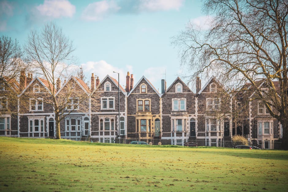 A row of Victorian-style terraced houses with dark brick facades, white window frames, and decorative architectural details, situated behind a well-maintained grassy area with a few leafless trees. The houses have gabled roofs and multiple chimneys, and are located on a street with a clear, partly cloudy sky overhead. In the foreground, the open lawn extends towards the houses, emphasizing the residential character of the area. This setting with traditional homes is often associated with property relocation and home moving services, supported by companies like [COMPANY_NAME] specializing in removals for house moves and furniture transport within Kentish Town. The scene captures the calm and orderly environment typical of a suburban neighbourhood ready for a home relocation or move, with the natural lighting highlighting the textures of the brickwork and the seasonal trees.