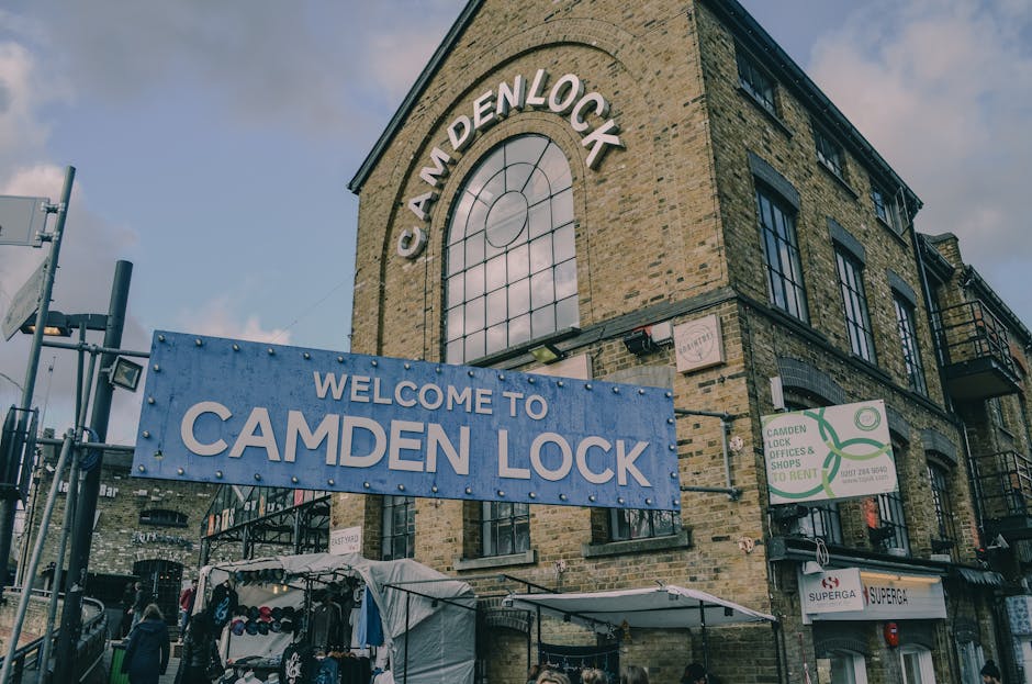 The image shows a three-story brick building with large black-framed windows, situated on a street corner in Camden Lock. A prominent blue sign with white lettering reading 'WELCOME TO CAMDEN LOCK' is suspended above the ground level, indicating the location. The building's façade features a large arched window near the top with the text 'CAMDEN LOCK' displayed in curved white letters. To the right, a green and white sign advertises 'Camden Lock Offices & Shops To Rent.' On the street level, there is a small outdoor market stall covered by a white canopy, selling various clothing items and accessories, with a few pedestrians nearby. The scene includes various equipment related to house or commercial move logistics, such as a trolley, plastic-wrapped furniture, or boxes, although they are not clearly visible in detail. The setting suggests an active area involved in residential or commercial relocations, with the building serving as a notable landmark in Camden Town, London. The lighting indicates daytime with a partly cloudy sky.