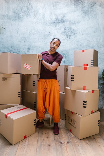 A person standing in an indoor space surrounded by neatly stacked cardboard boxes of various sizes, some sealed with red and black packing tape, ready for a home relocation or furniture transport. The individual is wearing a purple t-shirt and orange trousers, holding one of the boxes while looking at the camera. The background features a textured light blue wall and wooden flooring, with soft natural or artificial lighting illuminating the scene. The image depicts the packing and loading process common in house removals, as seen in professional moving services like those provided by removal companies in Kentish Town, emphasizing careful handling and organization during a house move.