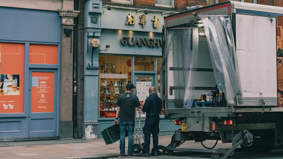 Two movers from [COMPANY_NAME] are preparing to load goods into a delivery truck outside a blue shopfront with Chinese characters and the word 'GUANGH' visible. One mover, wearing a black jacket and jeans, is holding a green plastic crate while the other, in a dark jacket and pants, is operating a hand truck. The truck is positioned on the pavement with its side open, revealing a plastic curtain and a space for loading. Nearby, cardboard boxes and packing materials are arranged on the ground, indicating a house relocation or furniture transport process. The scene is set during daytime with ample natural light, illustrating an organized moving operation supporting packing and moving services connected to the house removals process described on the Kentish Town loading bay rules page.