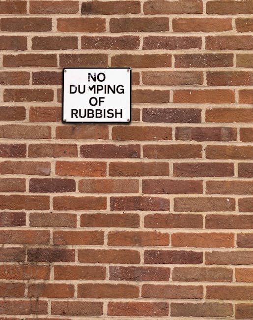 A white rectangular sign attached to a red brick wall with a black border and black text reading 'NO DUMPING OF RUBBISH.' The brick wall features a traditional running bond pattern with reddish-brown bricks and light grey mortar joints. The lighting is even, illuminating the textured surface of the bricks. This image relates to house removals and waste regulations that may be relevant to relocation services offered by companies like [COMPANY_NAME], emphasizing proper waste management during home relocation or furniture transport processes. The sign’s placement on the exterior wall indicates a regulation meant to prevent improper rubbish disposal, supporting environmentally responsible moving practices. The wall serves as a backdrop for clearly visible signage that conveys local rules or restrictions, useful for ensuring compliance during residential moves, packing, and loading activities involved in removals.