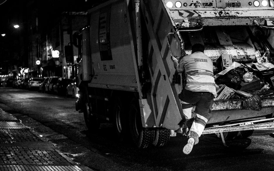 A sanitation worker dressed in a high-visibility uniform with reflective strips is seen climbing into the back of a large waste collection truck parked on a dimly lit urban street at night. The truck's rear compartment is open, revealing a variety of garbage bags, cardboard boxes, plastic containers, and other waste materials, some of which are loosely piled and spilling over the edges. The vehicle is positioned partially on the pavement with its rear elevated to facilitate loading, and the worker is using a hand to steady himself as he ascends. The street in the background shows parked cars, streetlights, and illuminated shop signs, indicating an active city environment at night. This scene captures the process of waste collection, relevant to urban housekeeping and municipal waste management, important considerations in home relocation or property clearing services provided by [COMPANY_NAME], within the scope of removals and moving logistics.