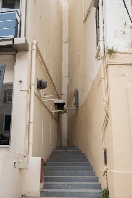 A narrow, steep outdoor staircase made of stone or concrete leads upward between tall, closely spaced residential buildings with white and beige facades. The buildings feature small, rectangular windo