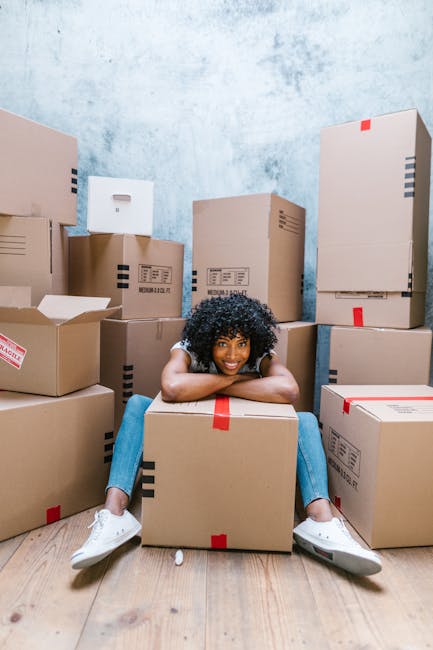 A man with curly hair and medium skin tone is seated on a wooden floor inside a room, surrounded by several cardboard moving boxes that are sealed with red packing tape. The boxes vary in size and are stacked beside him, some leaning against a beige cushioned chair. Behind him are two large, arched windows with white frames, allowing natural daylight to illuminate the space and offering a view of parked cars and a building across the street. To the right of the man, there is a tall potted plant with broad, dark green leaves. The room appears to be in the process of a home relocation, with the boxes ready for transport. The setting reflects a moving or packing situation typical of house removals, emphasizing the logistical aspect of furniture transport and packing during a house move, as part of professional services such as those offered by removal companies in Kentish Town.