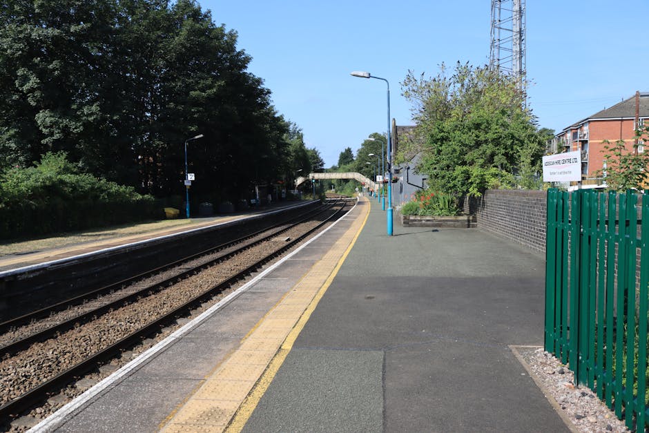 A view of a train station platform in daylight at Kentish Town with a clear blue sky. The platform has a yellow tactile paving strip along the edge and a smooth grey surface. A set of railway tracks runs parallel to the platform, with gravel bedding beneath the steel rails. On the right side, there is a green metal fence bordering the platform, and a small garden bed with plants and flowers next to a brick wall. Several blue-painted lampposts are positioned along the platform, and a few evenly spaced shelter structures are visible further along the platform. In the distance, a pedestrian bridge spans over the tracks, and nearby residential houses are partially visible behind trees. This scene illustrates a typical station environment suitable for house removals and furniture transport logistics, with no visible moving activity or objects being carried.