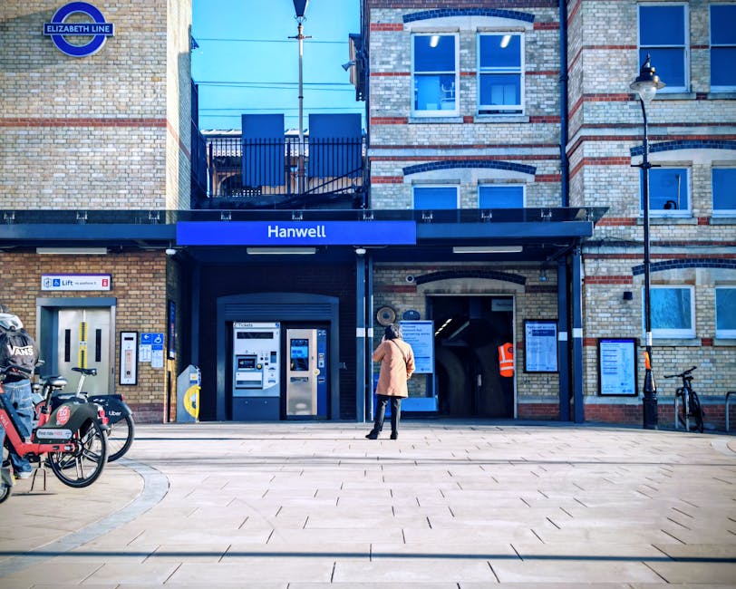 A person stands outside the entrance to Hanwell station, part of the Elizabeth Line, with a modern brick and concrete building featuring large blue signs, including the station name 'Hanwell' and the Elizabeth Line logo. The individual, dressed in a light brown coat and dark trousers, appears to be waiting or preparing for a home relocation, as there are bicycles and a Santander bike sharing station nearby. The station entrance is equipped with ticket machines, a lift for accessible entry, and electronic information boards mounted on the exterior wall. To the left, a bicycle rack contains two bikes, including a red Uber bike, and a black bicycle is parked near a lamppost on the sidewalk. The area is well-lit with daylight, and the open pavement provides space for loading and moving activities, reflecting the logistics involved in furniture transport and packing during house removals, coordinated by [COMPANY_NAME].