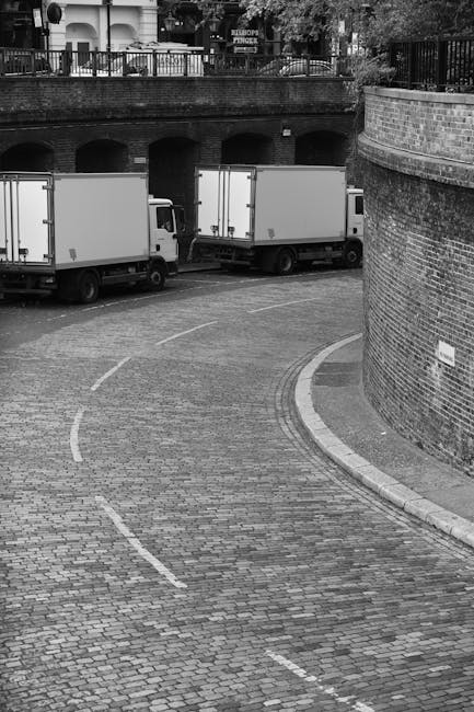 The image shows a curved cobblestone driveway or courtyard with two white box trucks parked side by side, likely used for home relocation or moving services. The trucks are positioned near a brick wall with an arched structure beneath a raised walkway or bridge, which features a metal railing and some trees or foliage in the background. The loading area is clear, with no visible activities happening at the moment, but the trucks are ready for loading or unloading furniture, boxes, or appliances. The scene is in black and white, highlighting the textured surface of the cobblestones, the brick wall, and the vehicles. This setting suggests a professional moving operation, possibly by [COMPANY_NAME], carrying out furniture transport or home packing and moving in Kentish Town. The overall environment appears organized and prepared for a swift relocation process, emphasizing the logistical aspects of same-day or last-minute removals. This image reflects the typical environment for local house removals, including vehicle positioning and the urban outdoor setting for efficient loading and unloading of household items.