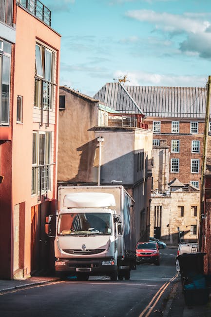 A narrow street in an urban area shows a moving van from uniserve.co.uk parked partly on a raised loading area, with a second larger truck behind it. The loading trucks are positioned directly on the road, which has railway tracks embedded in its surface. Two pedestrians, a woman and an older man, are crossing the street at a pedestrian crossing marked with white dashed lines. The woman, dressed in a dark jacket and jeans, is walking briskly while carrying a bag, and the older man, wearing a black coat and white shoes, is walking in the opposite direction. Surrounding buildings are tall, multi-storey structures made of concrete and brick, with windows and fire escape balconies, typical of a city environment. The scene captures a quiet moment during a home relocation or moving process, with the vehicles prepared for furniture transport and packing materials possibly unloaded from the trucks nearby. The lighting suggests an overcast day, providing even natural light across the scene, suitable for highlighting professional removals services like those offered by [COMPANY_NAME] in Kentish Town.