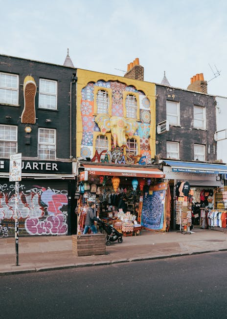 The image shows a row of commercial buildings on a street in Camden Road, Kentish Town, with a focus on a shop that has a colorful, decorative facade featuring a large yellow and orange elephant sculpture above its entrance. The shop displays various items, including textiles, clothing, and souvenirs, hanging outside the storefront. To the left, graffiti art is visible on a closed shutter, and to the right, there are additional shop displays with merchandise such as clothes and accessories. In front of the shops, the sidewalk is visible with a person pushing a stroller and another walking nearby. The buildings behind the shops are multi-storey, with some windows and chimneys on the roof, and the overall scene is lit by natural daylight. This street scene, typical of the area, could relate to residential or commercial property moves, as part of urban relocation or packing and moving activities in Kentish Town, as referenced on the service page for small flat removals.