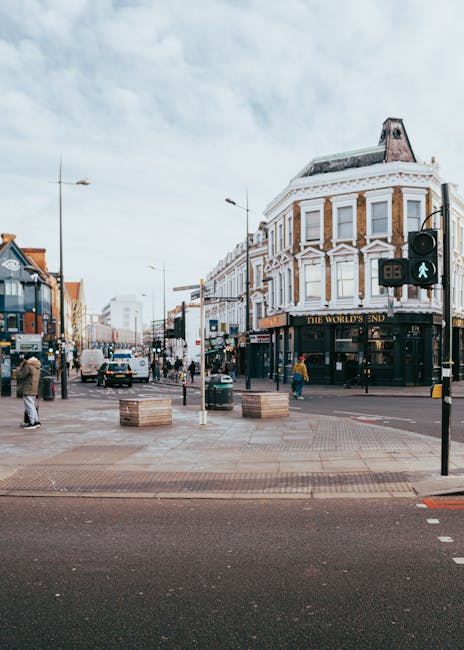 The image shows a row of commercial buildings on a street in Camden Road, Kentish Town, with a focus on a shop that has a colorful, decorative facade featuring a large yellow and orange elephant sculpture above its entrance. The shop displays various items, including textiles, clothing, and souvenirs, hanging outside the storefront. To the left, graffiti art is visible on a closed shutter, and to the right, there are additional shop displays with merchandise such as clothes and accessories. In front of the shops, the sidewalk is visible with a person pushing a stroller and another walking nearby. The buildings behind the shops are multi-storey, with some windows and chimneys on the roof, and the overall scene is lit by natural daylight. This street scene, typical of the area, could relate to residential or commercial property moves, as part of urban relocation or packing and moving activities in Kentish Town, as referenced on the service page for small flat removals.
