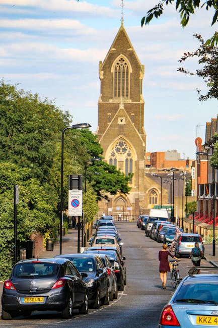 A row of parked vehicles, including small to medium-sized cars and a white van, line both sides of a narrow street leading towards a large historic church with a tall, pointed steeple and Gothic-style windows, set against a blue sky with scattered clouds. On the sidewalk, a person wearing a red coat is walking while pushing a bicycle, and another individual in casual attire is riding a bicycle past the parked cars. Lampposts and street signs are visible along the pavement, with green trees partially obscuring the lower parts of the church façade. The scene depicts a typical urban residential area in Kentish Town, suitable for house removals and furniture transport services by [COMPANY_NAME], highlighting the logistics involved in home relocation and packing within an organized city environment.